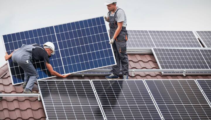 An image of a solar panel installation on the roof of a commercial building in Staten Island, NY. The installation team can be seen working on the roof, positioning the panels to capture maximum sunlight. The city skyline can be seen in the background, with clear blue skies and a few clouds. Tools, equipment, and safety gear are scattered on the roof.