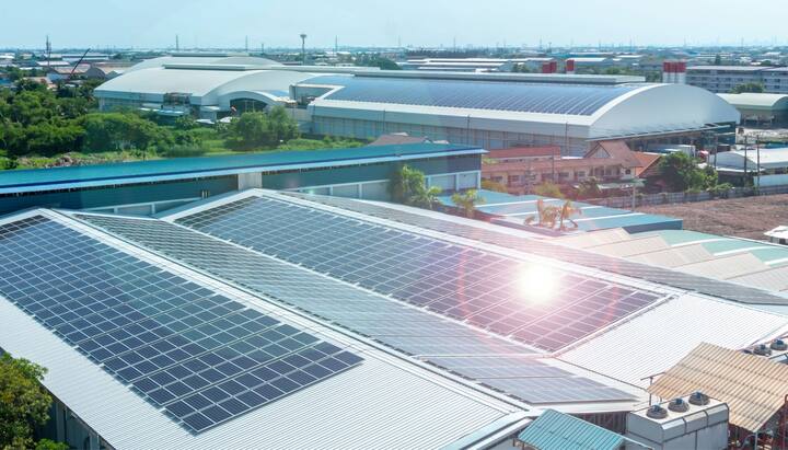 A commercial office building with solar panel installation on its rooftop in Staten Island, NY. The solar panels are positioned to capture maximum sunlight, with the city skyline visible in the background. The installation team can be seen working on the rooftop, arranging the panels and connecting them to the building's electrical system. The sky is cloudy with a hint of sunlight breaking through.
